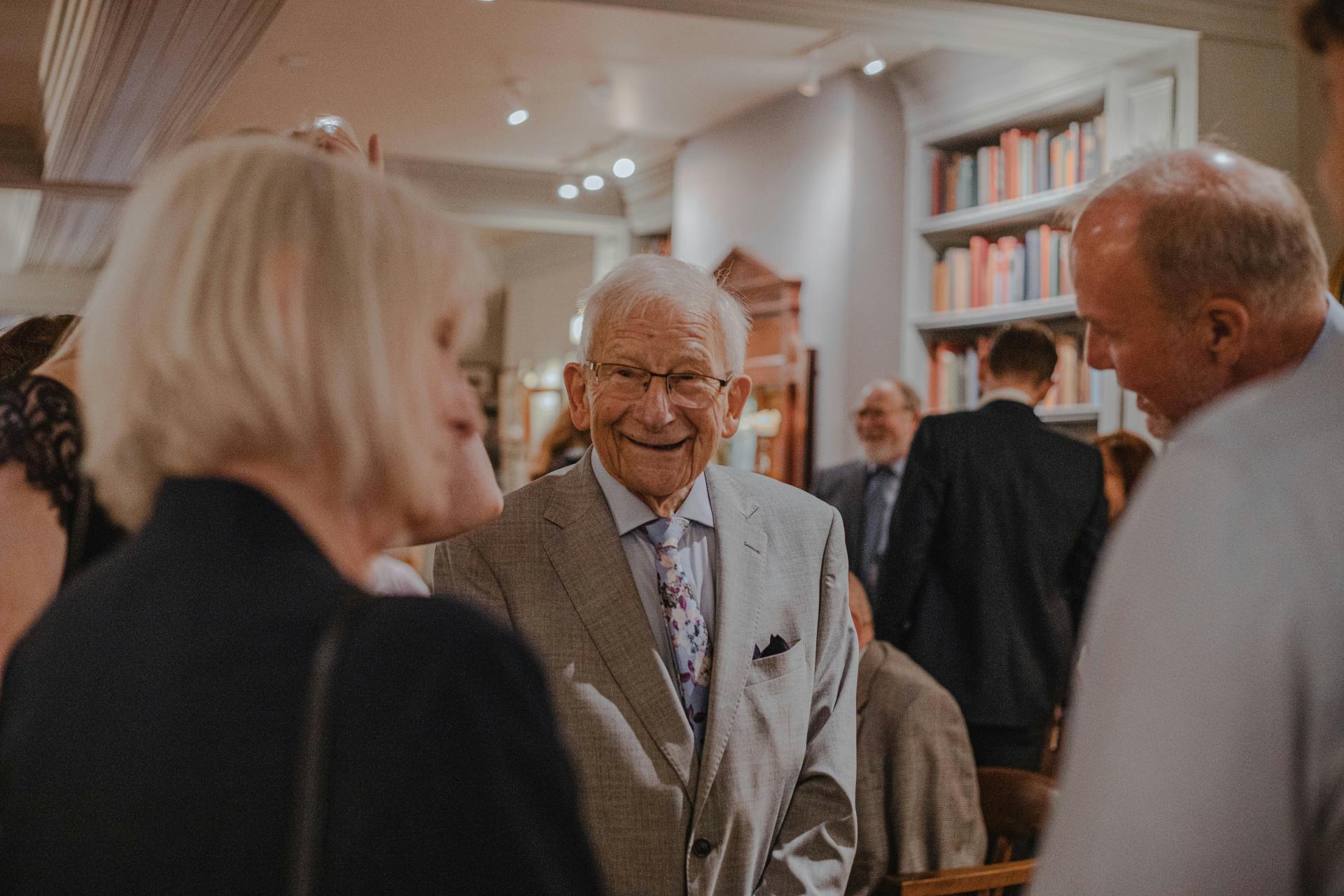 Hombre anciano de traje gris sonríe y habla con otras personas